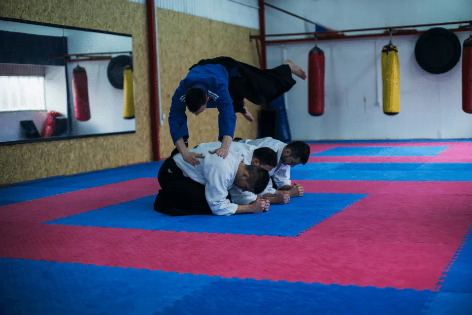 3 men in white kneeling on athletic mat, 1 man in blue somersaulting over them