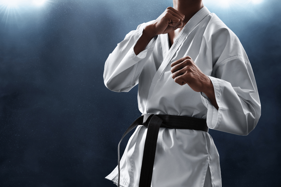 A man in martial arts gear, white robe, black belt, is posing on a dark background