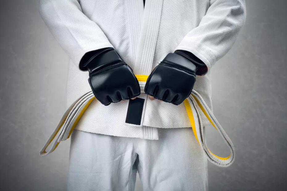 person in white karate robe, black gloves holding a hellow belt, equipment.